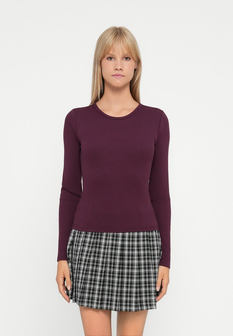 Burgunderfarbener, langärmeliger Rippenpullover, kombiniert mit einem schwarz-weißen karierten, plissierten Rock. Sanfte Textur und taillierte Silhouette.