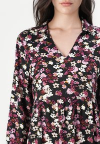 Robe noire à fleurs avec des manches longues, un col en V, un corps ajusté et une jupe évasée ornée de motifs de fleurs roses, blanches et violettes.