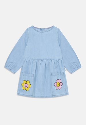 Lys blå denimkjole med lange ermer, en samlet midje og to frontlommer som har gule og rosa blomsterdesign med smilefjes.