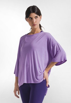 Vrouw met donker haar in een paardenstaart draagt een losse lavendelkleurige top en donkerpaarse broek, kijkt opzij met een hand op haar heup.