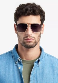 Occhiali da sole con forma rettangolare, lenti scure, montatura in metallo color oro e dettagli. Indossati da una persona in camicia di denim sopra una maglietta verde.