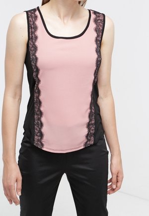 Kobieta w różowym topie bez rękawów z czarną koronkową lamówką na przednich panelach oraz w czarnych spodniach, stoi na jednolitym tle.