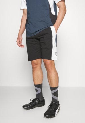 Homme portant un short de sport noir et blanc, des chaussettes noires et grises, des crampons noirs, et une chemise bleue et blanche à manches courtes, debout avec une main dans la poche.