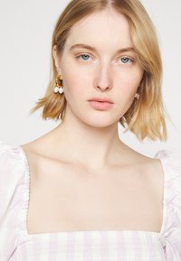 Modelo com cabelo loiro curto a usar um vestido lilás claro com mangas bufantes. Acompanhada por brincos de pérolas douradas e brancas.