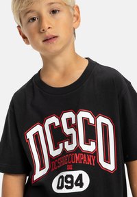 Čierne bavlnené tričko s výrazným červeným a bielym grafickým textom "DC Shoe Company" a "094," s klasickým dizajnom okrúhleho výstrihu.