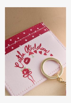 Portfel na klucze w kolorze białym i czerwonym z kropkami. Posiada kwiatowy wzór, napis oraz przymocowany złocisty metalowy brelok.