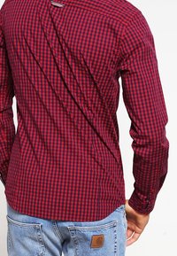 Man in een rood en zwart geruit overhemd met lange mouwen en lichtblauwe jeans, van achteren en tot aan de taille afgebeeld.