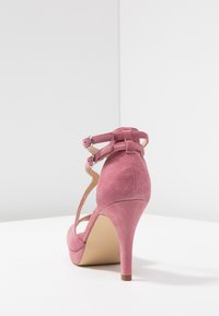 Zapato de tacón alto de ante rosa con tiras en el tobillo y punta afilada. Presenta una textura suave y un tacón esbelto con base circular.