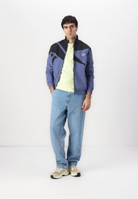 Blå og sort lynlåsjakke med geometriske designs, parret med en lys grøn t-shirt, løse lyseblå jeans og beige sneakers.