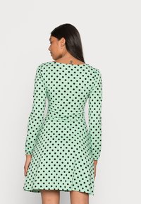 Vestido verde menta com mangas longas, apresentando bolinhas pretas. A saia tem um leve efeito evasê, com um decote redondo e cintura ajustada.