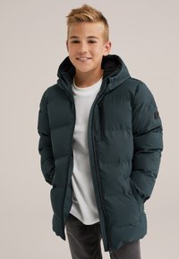 Temno zelena podložena jakna s kapuco, sprednjim zadrgo, stranskimi žepi in diskretno zakrpano oznako. Nosi se čez belo majico in sivo jeans hlače.