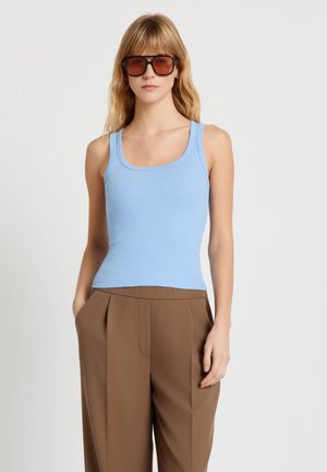 Donna con occhiali da sole marroni oversize, top smanicato azzurro chiaro a coste e pantaloni marroni a gamba larga e vita alta, in piedi con le mani in tasca.