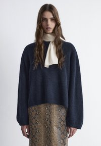 Navy Oversize-Pullover mit langen Ärmeln, aus weichem Material, kombiniert mit einem beigen Schal und einem braunen Rock im Schlangenmuster.