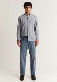 Camicia button-up grigia chiara con taschino sul petto, indossata con jeans blu a gamba dritta e scarpe nere, in piedi davanti a uno sfondo semplice.