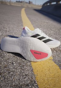 Weiße Laufschuhe mit schwarzen Streifen, auf Asphalt platziert, mit der strukturierten Sohle und einem roten Continental-Logo-Patch.