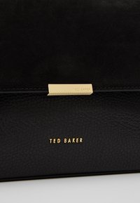 Černá kožená kabelka s texturovaným povrchem, zlatými kovovými doplňky a embosovaným logem. Jednoduchý, strukturovaný design s klopou.