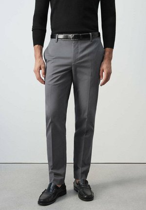 Hombre con pantalones grises ajustados, cinturón negro, mocasines negros y una camiseta negra de manga larga, de pie sobre un suelo gris.