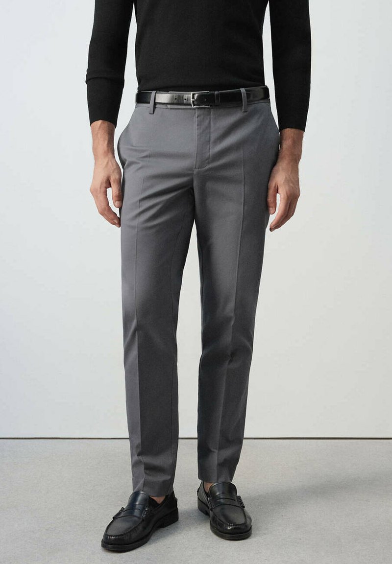 Homme portant un pantalon gris ajusté avec une ceinture noire, des mocassins noirs et un haut noir à manches longues, debout sur un sol gris.