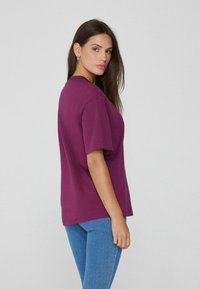 Camiseta de manga corta y cuello redondo en un profundo color púrpura, fabricada en algodón. Cuenta con un corte relajado y una textura suave, combinada con unos vaqueros ajustados azules.