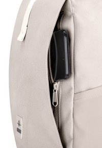 Sac à dos en tissu beige avec une poche latérale zippée partiellement ouverte montrant un smartphone noir à l’intérieur.