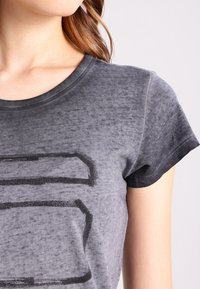 Grå bomullströja med rundad halsringning, med ett urblekt mönster och korta ärmar. Texturen verkar mjuk med en lätt sliten finish.