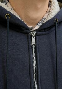 Granatowy rozpinany hoodie z miękkim, polarowym wnętrzem. Posiada srebrny zamek błyskawiczny, czarne sznurki i klapki w kratkę widoczne spod spodu.