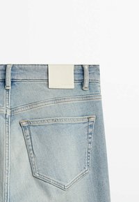Jean en denim délavé clair avec une conception standard de poche arrière et une étiquette rectangulaire en cuir blanc à la ceinture. Détails de coutures visibles.