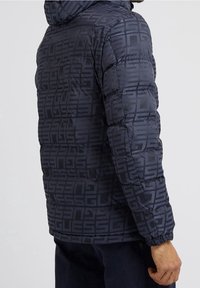 Marine blauwe pufferjas met een herhaald 'GUESS'-logo patroon, voorzien van een capuchon, een doorgestikt ontwerp, elastische manchetten en een gladde, glanzende textuur.