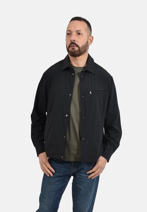 Uomo con barba corta che indossa una giacca nera abbottonata sopra una camicia verde oliva e jeans blu, in piedi con le mani vicino alle tasche.