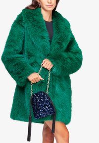 Abrigo de peluche verde con grandes solapas y mangas largas. Sosteniendo un bolso de lentejuelas azul marino con borlas negras y cadena.