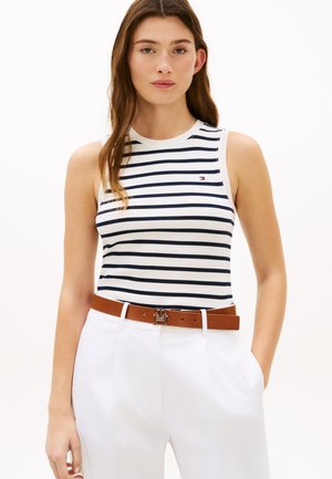 Femme portant un haut blanc sans manches avec des rayures bleu marine, un pantalon blanc et une ceinture marron avec une boucle métallique, debout avec une main dans la poche.