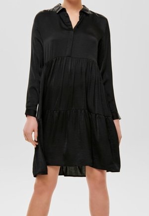 Zwarte jurk met lange mouwen, kraag en gelaagde rok. Gemaakt van gladde stof met een subtiele glans. Boven de knie lengte.
