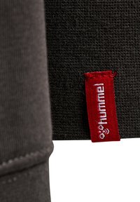 Nahaufnahme eines schwarzen Strickstoffs mit einem roten gewebten Etikett mit weißem „hummel“-Text und Logo, das am Rand befestigt ist.