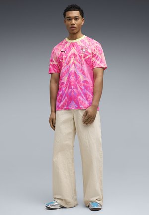 Młody mężczyzna stoi, ubrany w różową wzorzystą koszulkę piłkarską Portugalii, luźne beżowe spodnie i niebiesko-białe trampki na neutralnym tle.