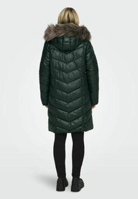 Manteau d'hiver vert rembourré avec un motif en chevrons, capuche doublée de fourrure et design mi-long. Porté avec un pantalon noir et des bottes noires imposantes.