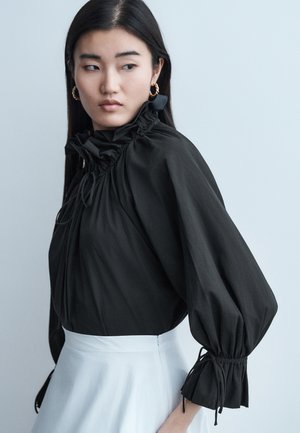 Mladá žena s dlhými tmavými vlasmi má na sebe čiernu volánovú blúzku s nazbieranými rukávmi a bielu sukňu, pozerá sa cez rameno.