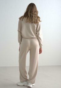 Beige Lounge-Set aus weichem Stoff, bestehend aus einem lässig geschnittenen Oberteil mit langen Ärmeln und passenden, leger geschnittenen Hosen; getragen mit weißen Sneakern.