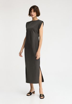 Mujer con vestido midi gris oscuro sin mangas con abertura lateral y sandalias negras de tacón, de pie frente a un fondo blanco.
