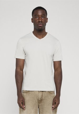 Ung sort mand står, iført en simpel hvid V-hals t-shirt med korte ærmer og beige denim bukser mod en ensfarvet grå baggrund.