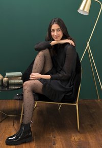 Black dress with ruffled hem, sheer polka dot tights, glossy black ankle boots, seated in a gold-accented chair against a green backdrop.