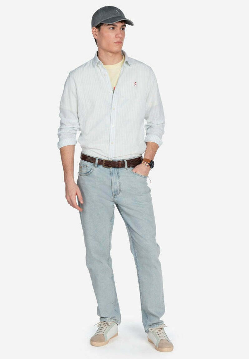 Camisa de botón de rayas azul claro, jeans azul claro, gorra gris, cinturón marrón y zapatillas blancas con acentos en beige. Manos en los bolsillos, pose de pie.