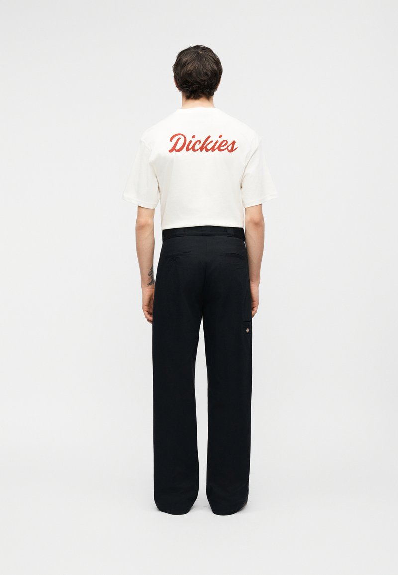 Homme debout de dos portant un t-shirt blanc « Dickies » et un pantalon noir à jambes larges sur un fond uni.