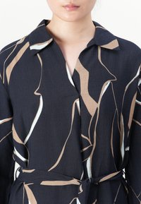 Femme portant une robe chemise noire avec un motif de lignes abstraites beige et blanches et une ceinture en tissu nouée à la taille.