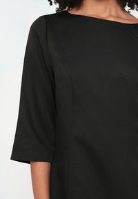 Μαύρο φόρεμα με λαιμόκοψη σκάφης, μανίκια τριών τετάρτων και λεία υφή. Διαθέτει διακριτική ραφή κατά μήκος του μπούστου.