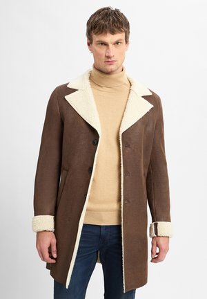 Brown suede coat with a white shearling collar and cuffs, featuring a button front and a tailored fit. Worn over a beige turtleneck.