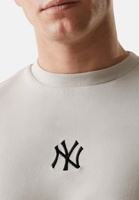 Felpa grigia chiara con scollo a girocollo, con un logo NY ricamato di colore nero sul petto. Tessuto liscio e design aderente.