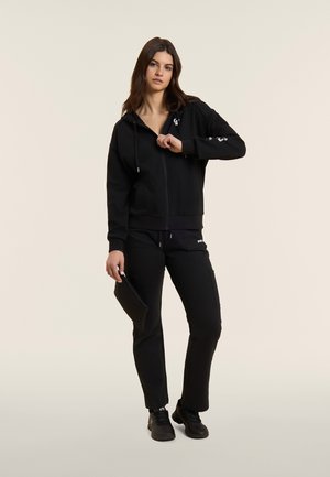 Giovane donna in piedi che tiene una pochette nera, indossa una felpa con zip nera, pantaloni cargo neri e sneakers nere su sfondo beige.