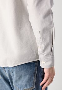 Lys grå langermet skjorte laget av teksturert stoff, med en knappelukking på mansjetten og en rett kant, sammen med blå denimjeans.