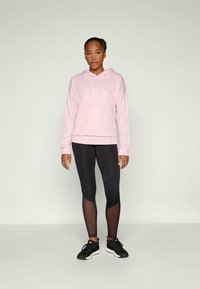 Kvinna som står i en ljusrosa hoodie, svarta leggings med meshpaneler och svarta träningsskor mot en enkel ljus bakgrund.
