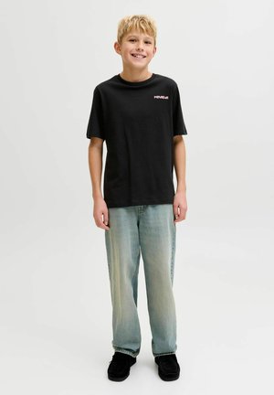 Niño sonriente de pie, vistiendo una camiseta negra con diseño gráfico, jeans azul claro y zapatos negros sobre un fondo blanco liso.